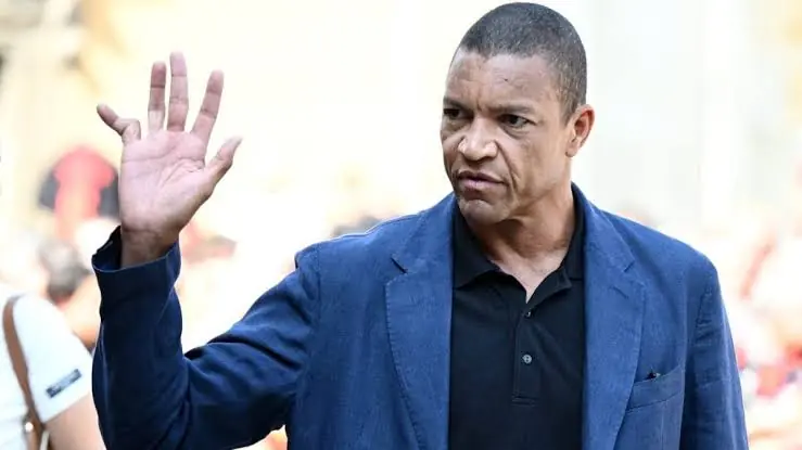 DIDA - THEWILL NEWS MEDIA Man in a blue blazer and black polo raises his right hand in a wave, outdoors, with a focused expression.
