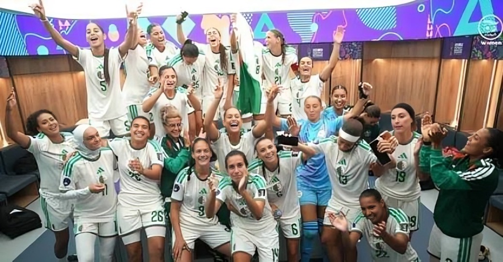 Algeria women's national team - THEWILL NEWS MEDIA Group of female players in white and green uniforms celebrate in a locker room, arms raised and smiling, with a blue goalie standing center and a colorful banner in the background.