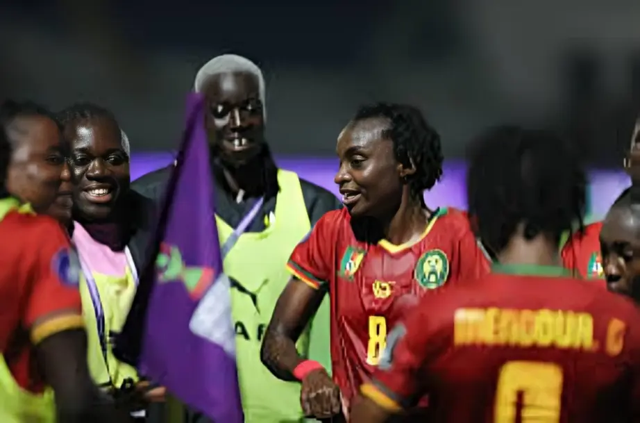 Cameroon women's national team celebrating - THEWILL NEWS MEDIA Group of soccer players in red uniforms celebrating on the field, with a purple flag/banner in the background.