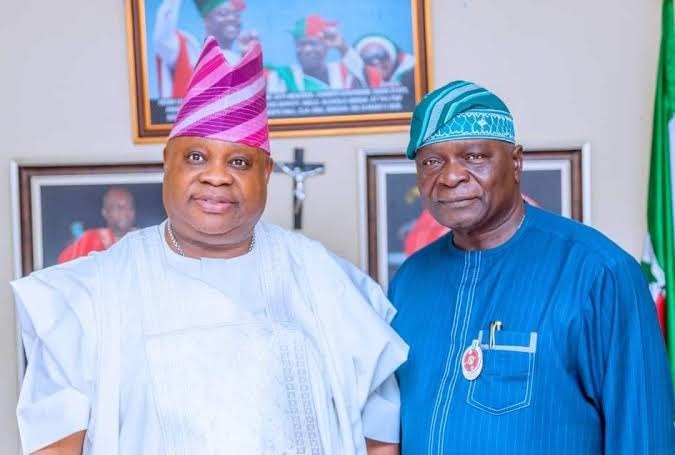 Adeleke and Oyinlola - THEWILL NEWS MEDIA Two Nigerian men in traditional attire posing indoors; one in a white agbada with a pink turban and the other in blue with a teal headwrap, framed photos on the wall behind them.