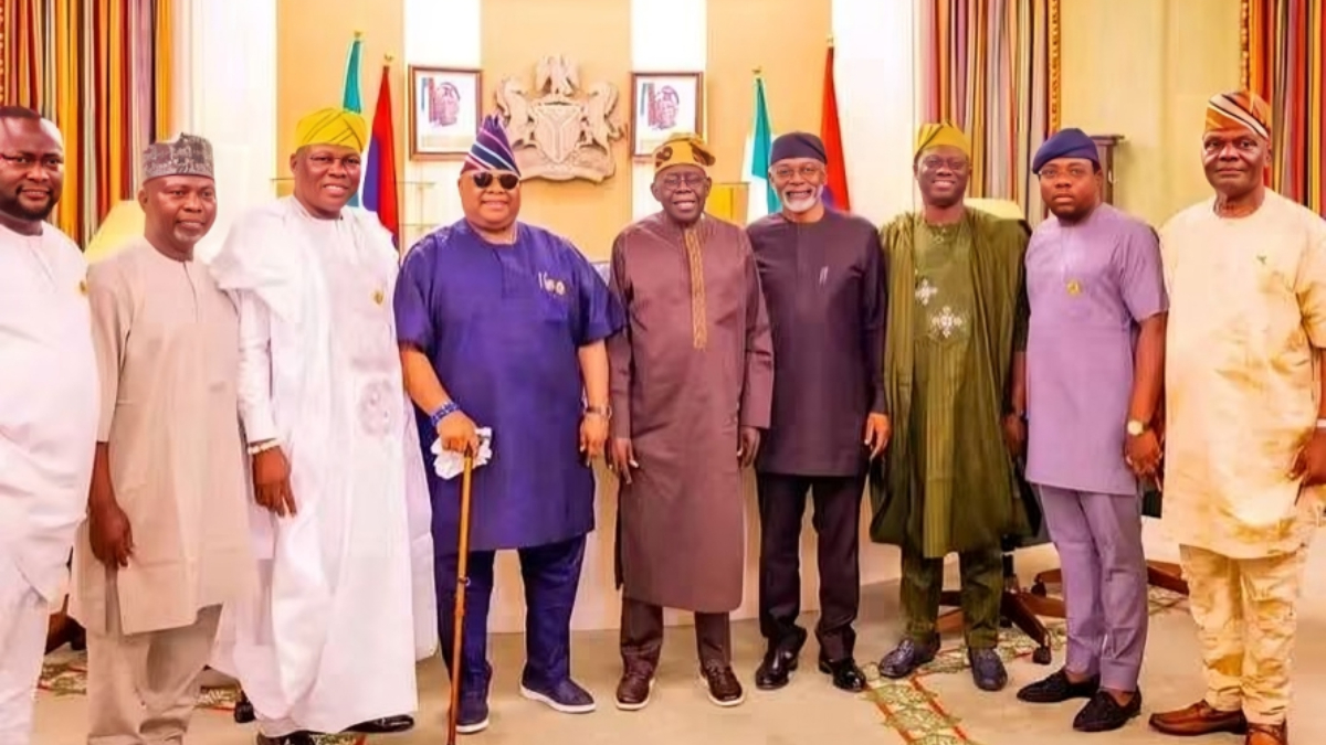 Adeleke in Aso Villa - THEWILL NEWS MEDIA Ten men in colorful traditional Nigerian attire pose in a formal room with flags and a coats‑of‑arms crest in the background, looking at the camera.
