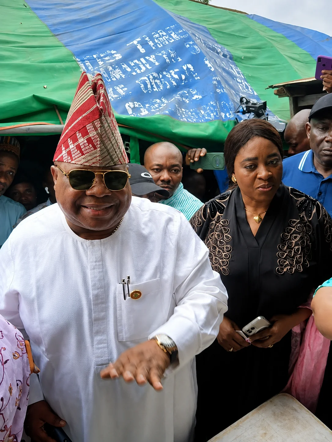 Adeleke votes - THEWILL NEWS MEDIA Smiling man in a white traditional outfit and red patterned hat wearing sunglasses, surrounded by a crowd under blue and green tarps.