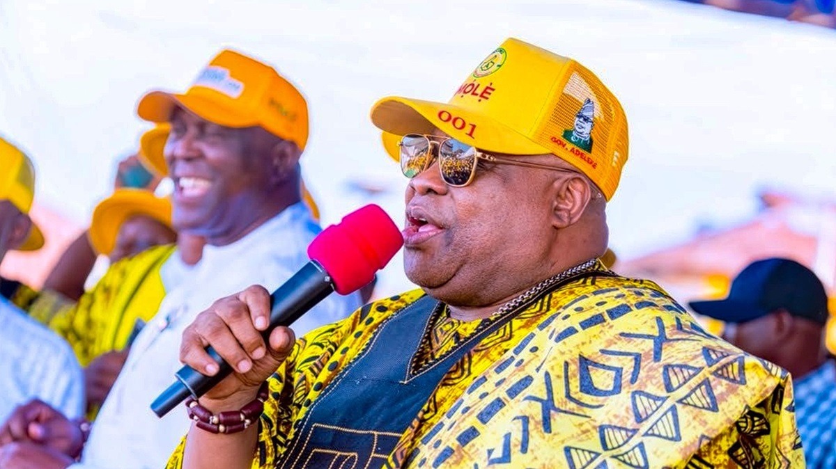 Ademola Adeleke - THEWILL NEWS MEDIA Man in a yellow patterned shirt and matching cap speaks into a red handheld microphone at an outdoor event, with supporters in yellow in the background.