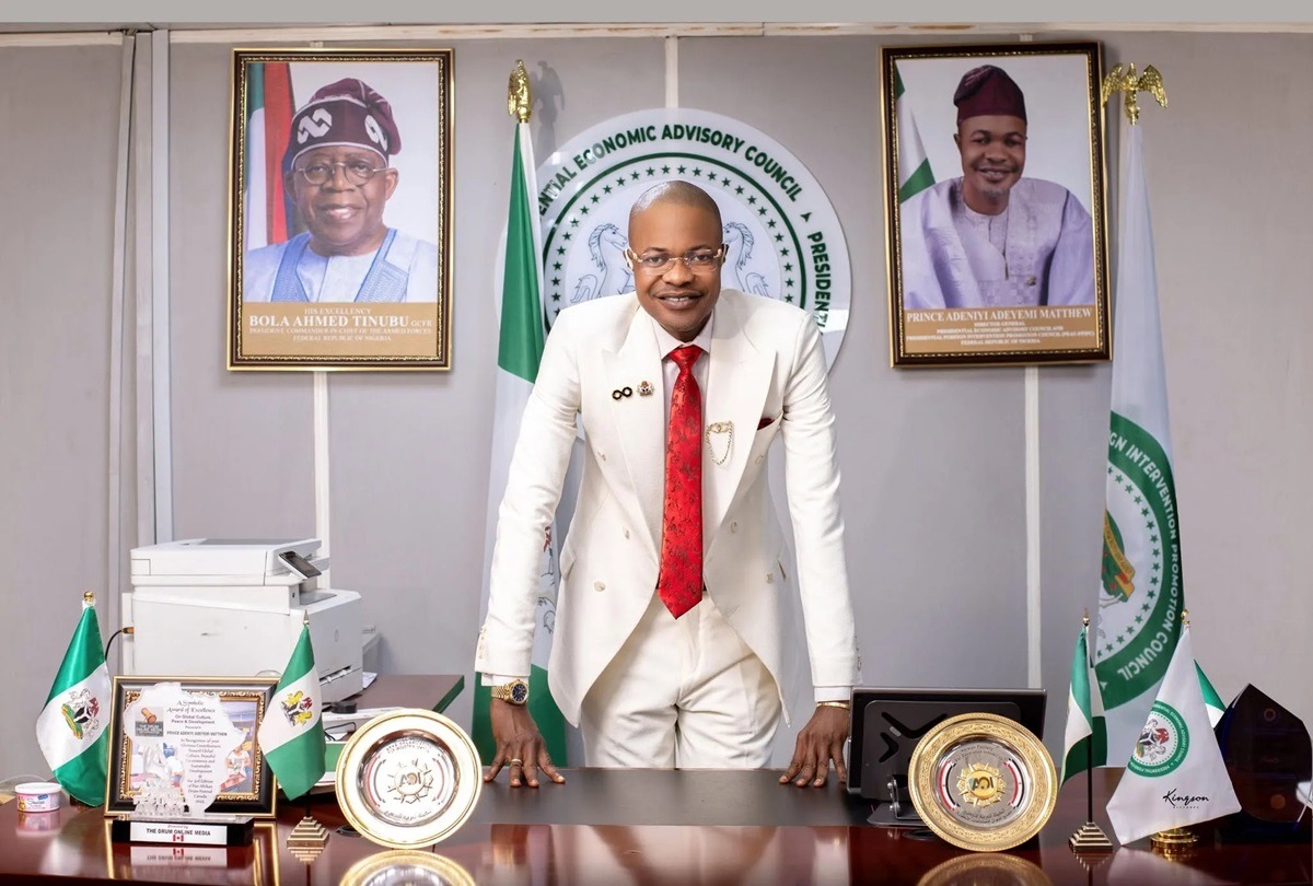 Adeniyi Adeyemi - THEWILL NEWS MEDIA Man in a white suit and red tie leans on a polished desk in a formal office, with Nigerian flags and framed portraits behind him.