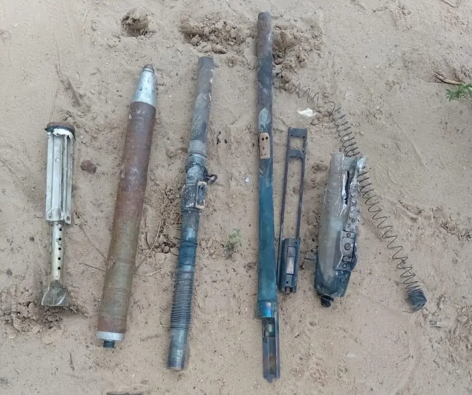 Amunitions - THEWILL NEWS MEDIA Row of rusted military ordnance components laid out on sandy ground, including cylindrical tubes and a coiled spring.