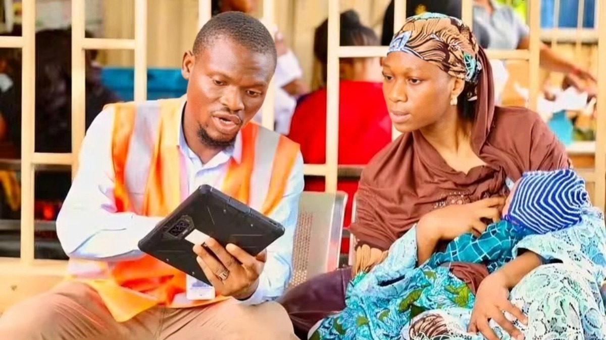 An NPC official guides a nursing mother - THEWILL NEWS MEDIA Man in a high‑vis vest showing a tablet to a mother holding a wrapped baby in colorful attire.