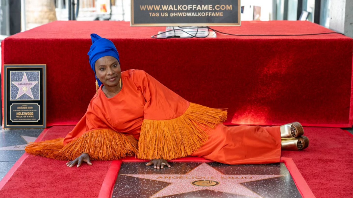 Angélique Kidjo - THEWILL NEWS MEDIA Woman in a vibrant orange dress with fringed sleeves and a blue headwrap posing on a Hollywood Walk of Fame star on a red carpet.