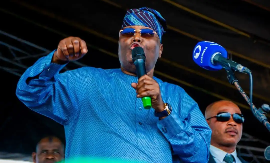 Atiku Abubakar - THEWILL NEWS MEDIA Man in a blue traditional outfit and head wrap speaks into a handheld microphone on stage, wearing sunglasses.