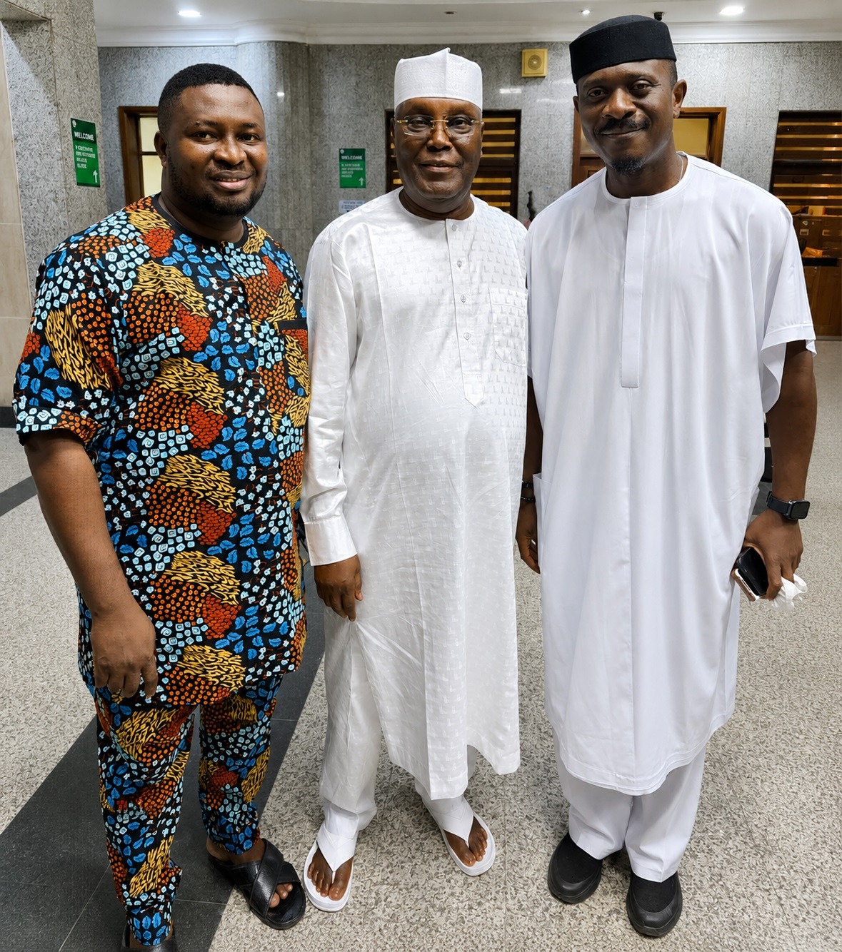 Atiku Seeks Court-Ordered Disqualification Over Certificate Allegations - THEWILL NEWS MEDIA Three men in traditional African attire pose together in a marble hallway; the left wears a colorful patterned outfit, the center a white agbada with a white cap, and the right a white tunic with a black cap.