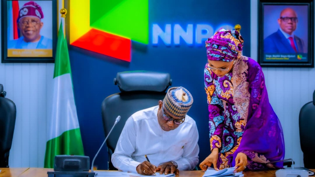 Bayo Ojulari - THEWILL NEWS MEDIA Man in white traditional attire signs a document at a table while a woman in purple traditional dress assists, with a Nigerian flag and NNPC backdrop behind them.