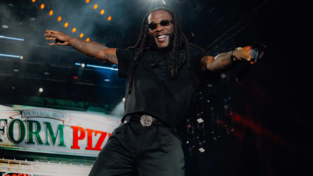 Burna Boy - THEWILL NEWS MEDIA Male performer on stage with arms outstretched, wearing black and sunglasses, under bright stage lights with a signage backdrop behind him.