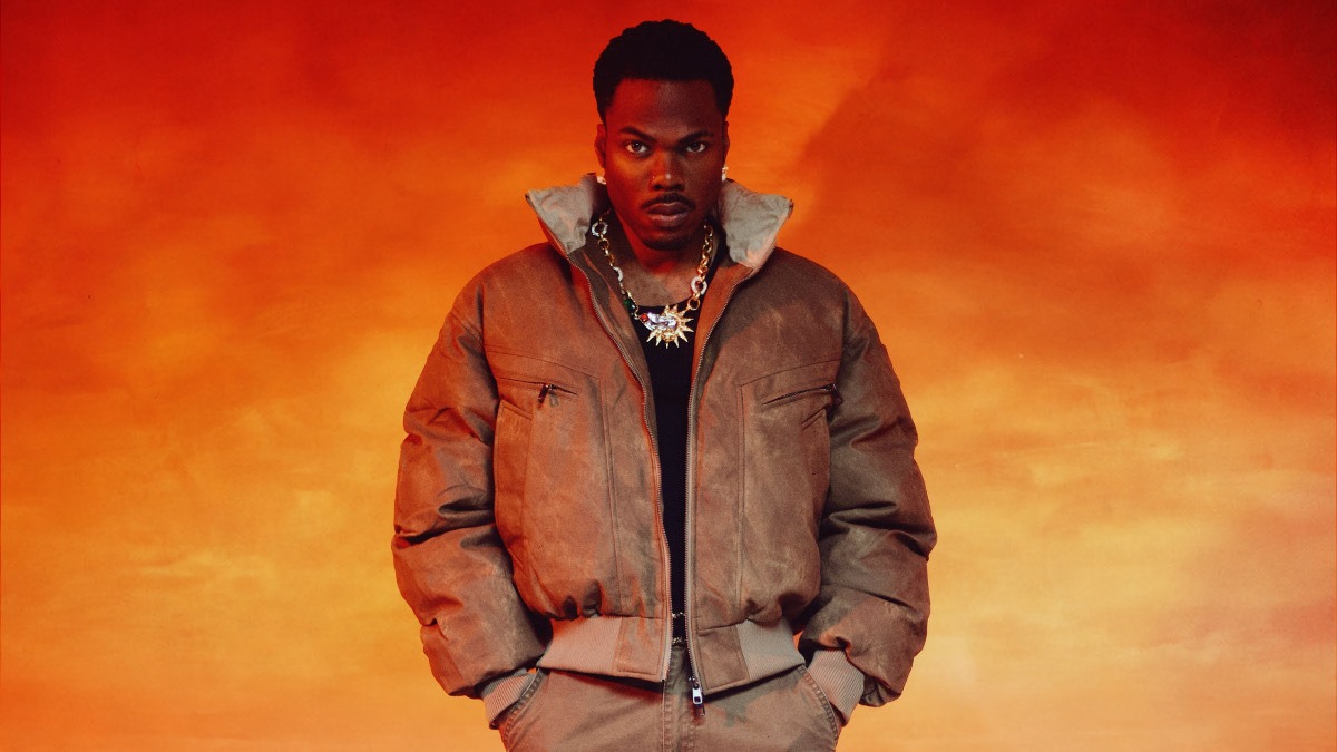 CKay - THEWILL NEWS MEDIA Man standing against an orange gradient background, wearing a brown puffer jacket and chunky gold chain, looking at the camera with a serious expression