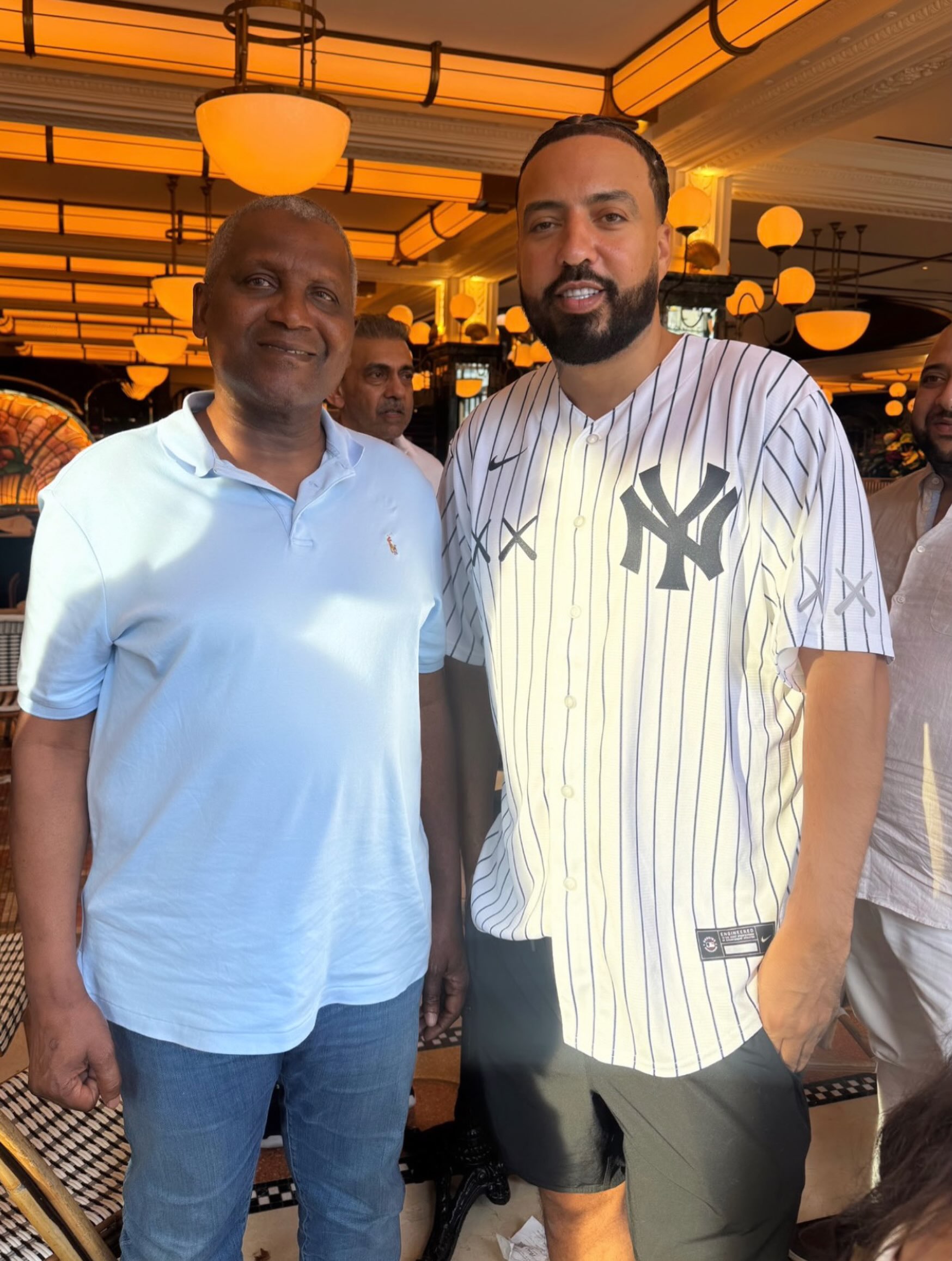 Dangote French Montana - THEWILL NEWS MEDIA Two men pose together indoors under warm orange lighting; one in a light blue polo shirt and the other in a white pinstripe Yankees jersey and dark shorts.