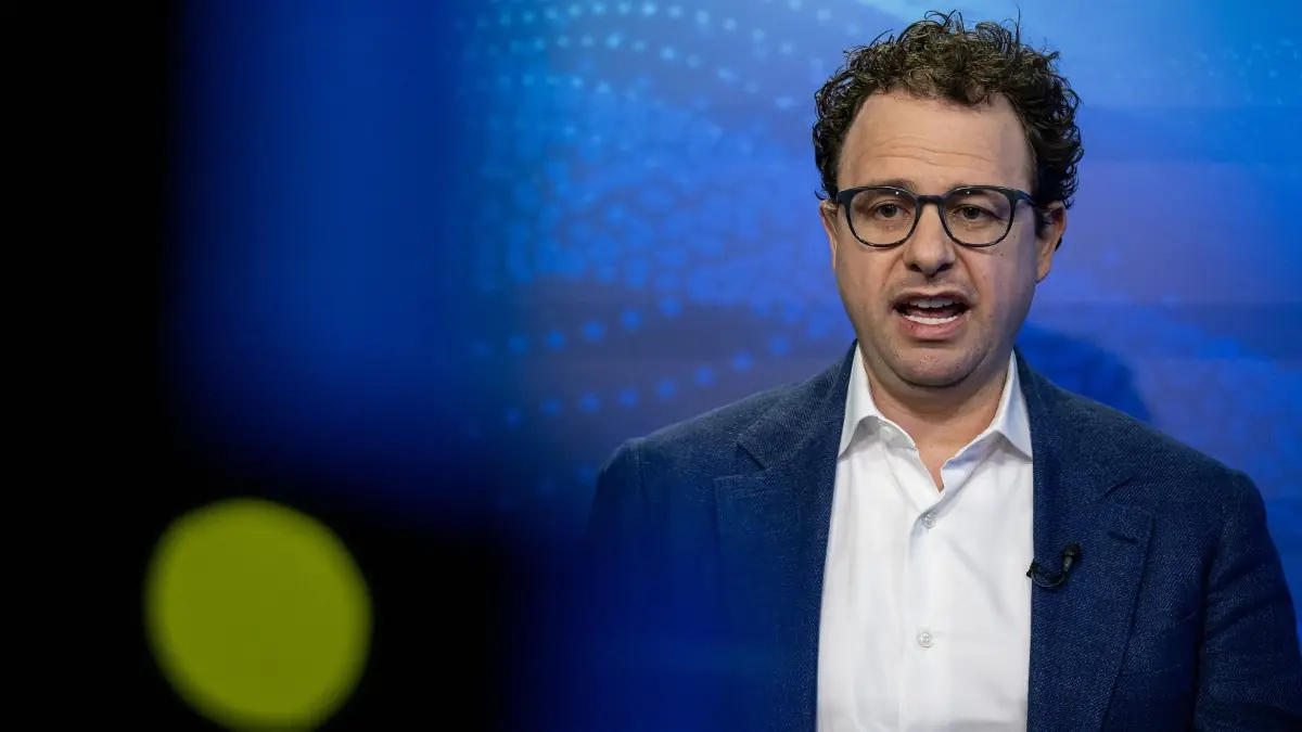 Dario Amodei - THEWILL NEWS MEDIA Man in a white shirt and navy blazer speaks on stage, wearing glasses, with a blue patterned backdrop.