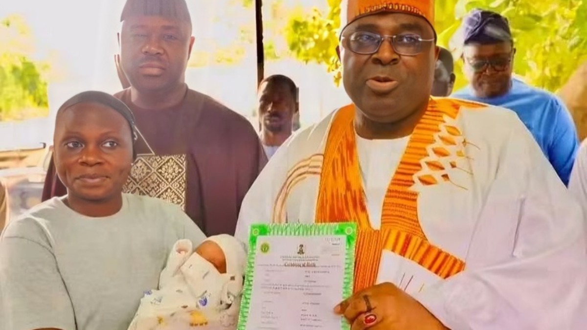 Dr Aminu Yusuf right presents a digital birth certificate to a mother - THEWILL NEWS MEDIA Woman holds a newborn swaddled in white, while a man in white and orange attire shows a green-bordered certificate to the camera, with onlookers in background.