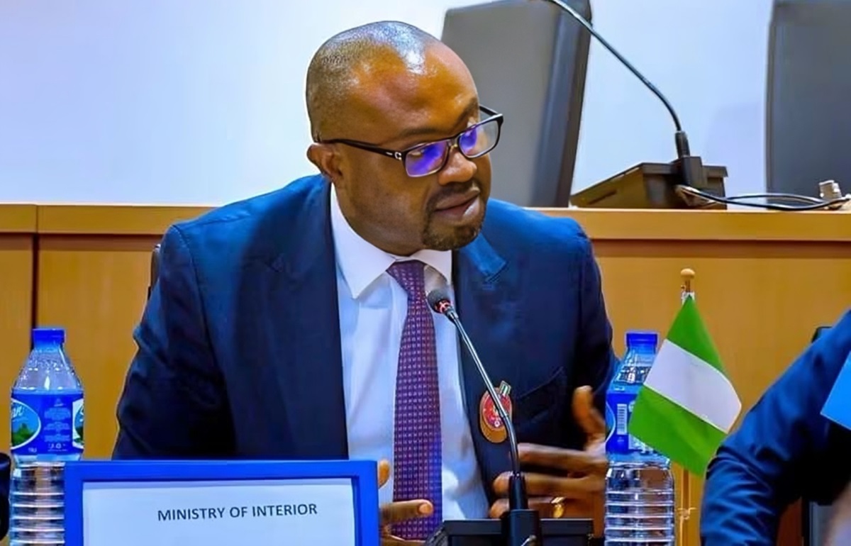 Man in a blue suit and glasses speaks into a microphone at a formal meeting; sign on the table reads'MINISTRY OF INTERIOR'.