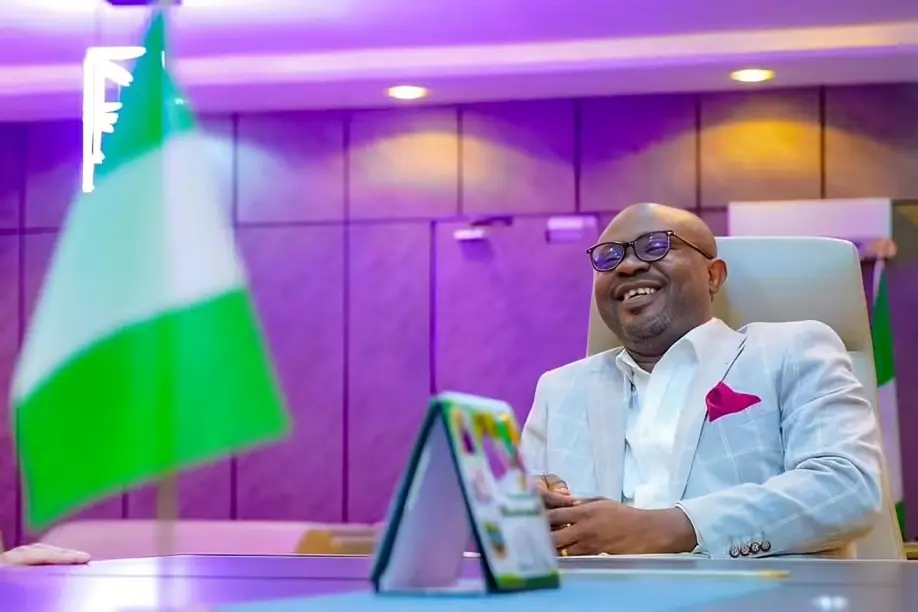 Dr Olubunmi Tunji-Ojo - THEWILL NEWS MEDIA Smiling man in a light blue blazer and glasses sits at a desk in a purple-lit conference room, with a green-and-white flag nearby and a tablet on the desk.