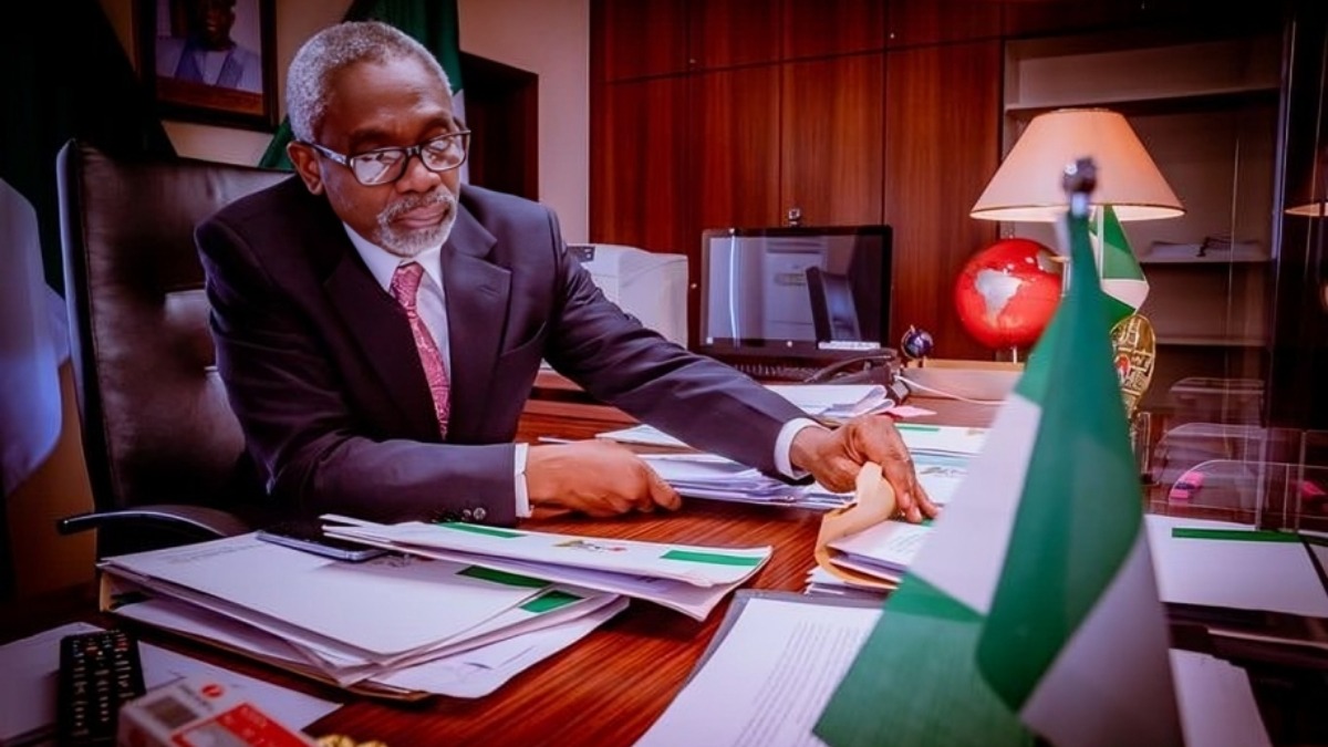 Femi Gbajabiamila - THEWILL NEWS MEDIA A suited man with glasses sits at a cluttered wooden desk, sorting through papers with a Nigerian flag in the foreground.