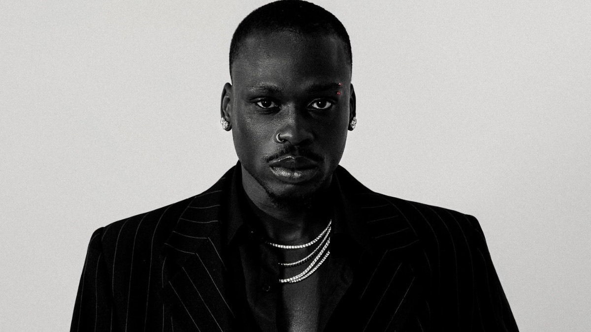 Fireboy - THEWILL NEWS MEDIA Black-and-white portrait of a man in a pinstripe suit with layered silver necklaces and a nose ring, looking at the camera.
