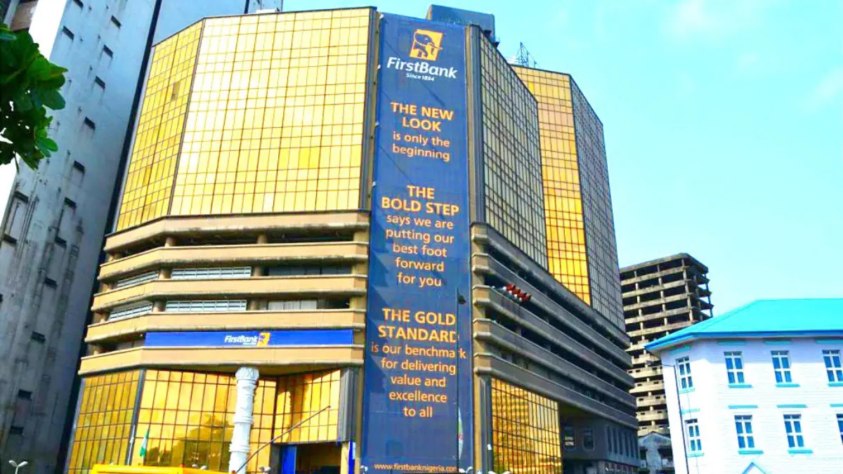 Firstbank - THEWILL NEWS MEDIA Facade of a modern bank building with large yellow glass panels and a vertical blue banner reading 'FirstBank' and promotional text.