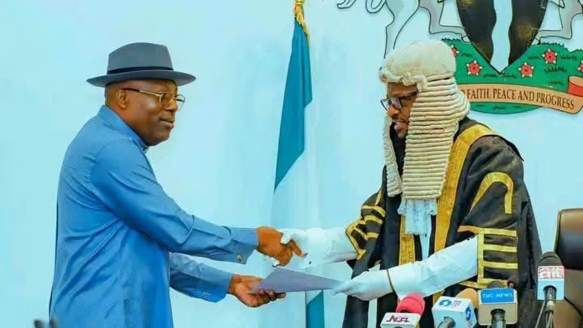 Fubara - THEWILL NEWS MEDIA Two men shake hands as one hands a document to a man in ceremonial judicial robes with a white wig; press microphones sit in front, a Nigerian flag visible in the background.