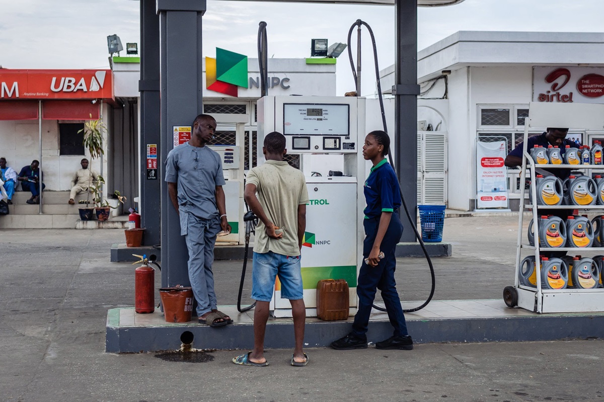 Fuel attendant at a petrol pump - THEWILL NEWS MEDIA Three men stand near a gas pump at a station; a uniformed attendant chats with a customer in a beige shirt while another man watches nearby.