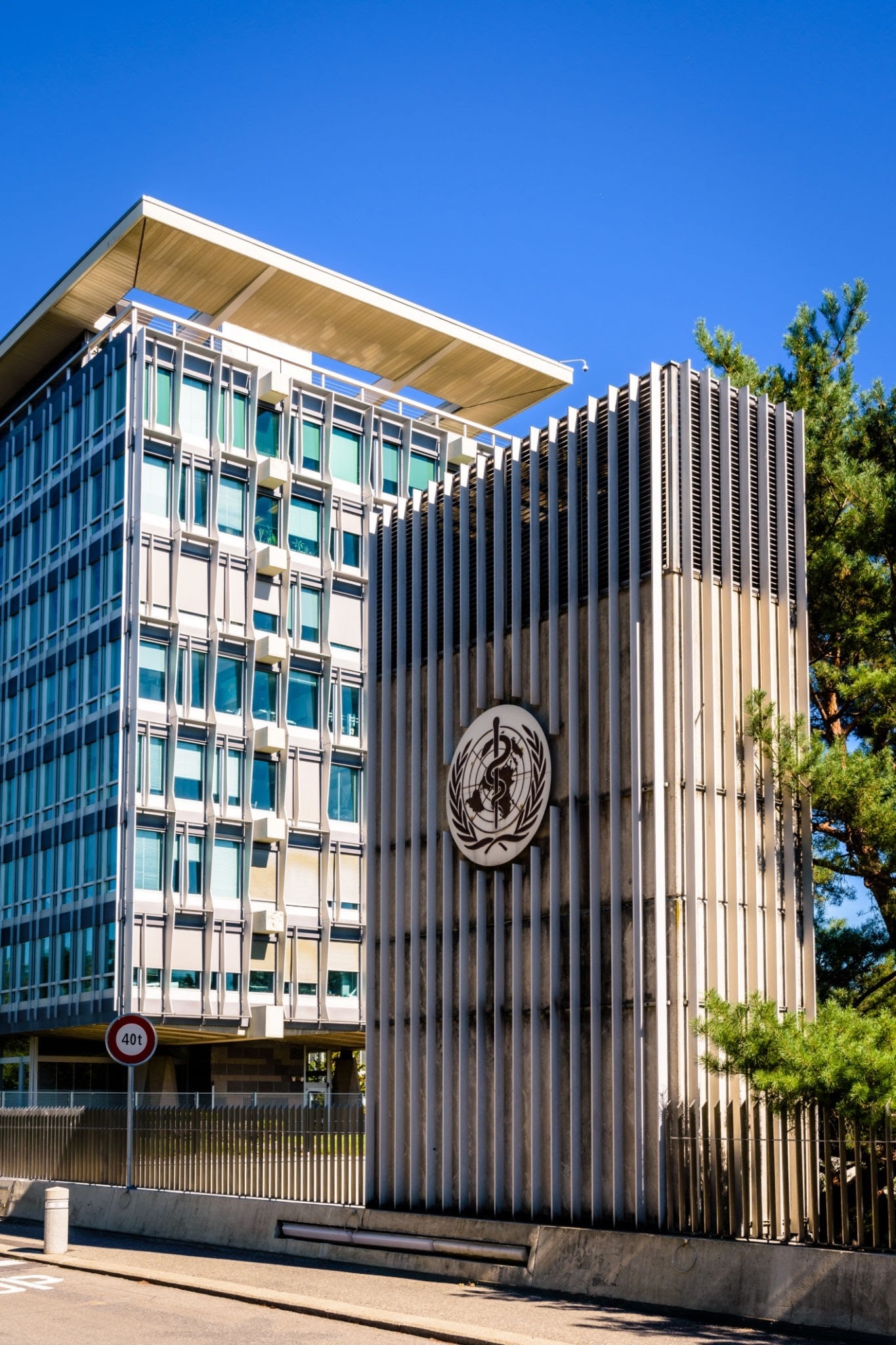 Geneva Hospital - THEWILL NEWS MEDIA Modern office building with glass windows and white vertical louvers, under a bright blue sky; a circular emblem is mounted on the exterior wall.