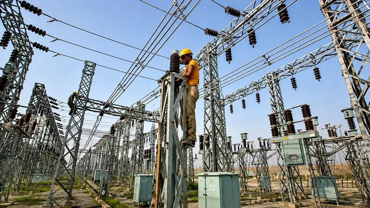 Grid - Substation - Electricity - THEWILL NEWS MEDIA Two workers in yellow helmets perform maintenance on high‑voltage electrical equipment in a metal lattice substation, with many power lines overhead.