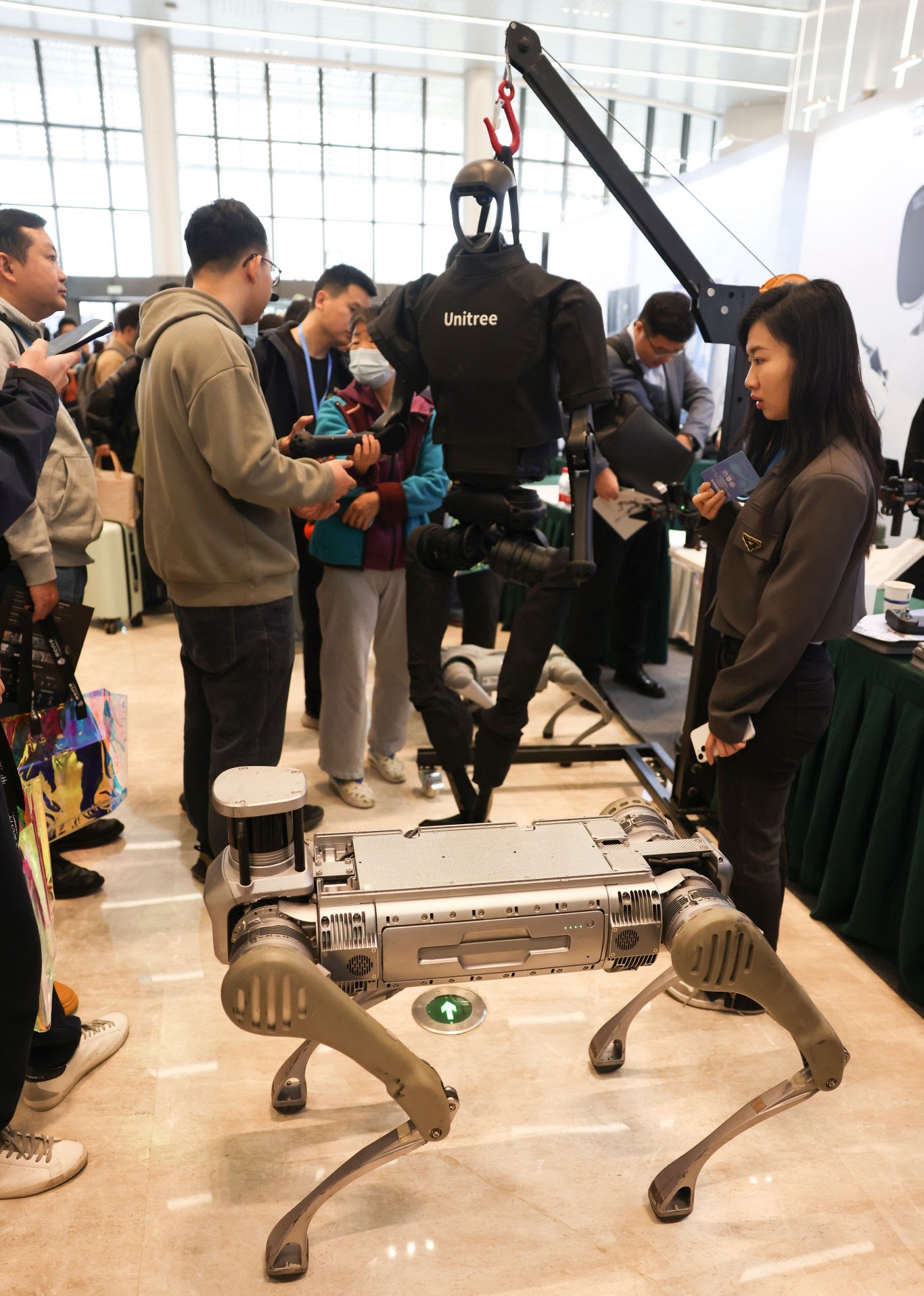 Humanoid Robot - THEWILL NEWS MEDIA Crowd at a tech expo inspecting a Unitree humanoid robot mounted on a mechanical crane.