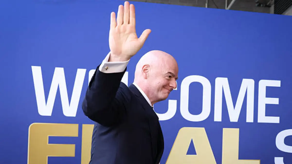 Bald man in a dark suit waves with his right hand in front of a blue backdrop that reads'WELCOME'.