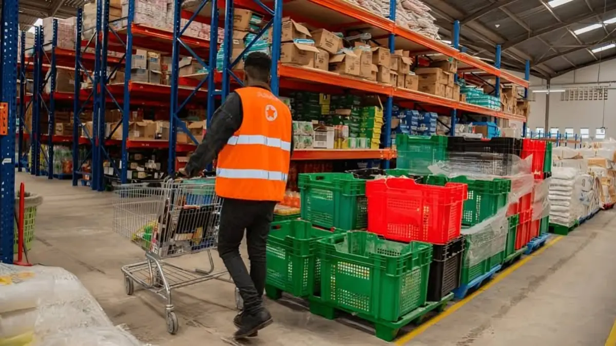 JUMIA - THEWILL NEWS MEDIA Worker in an orange safety vest pushes a shopping cart through a warehouse aisle with tall shelves and stacked crates.