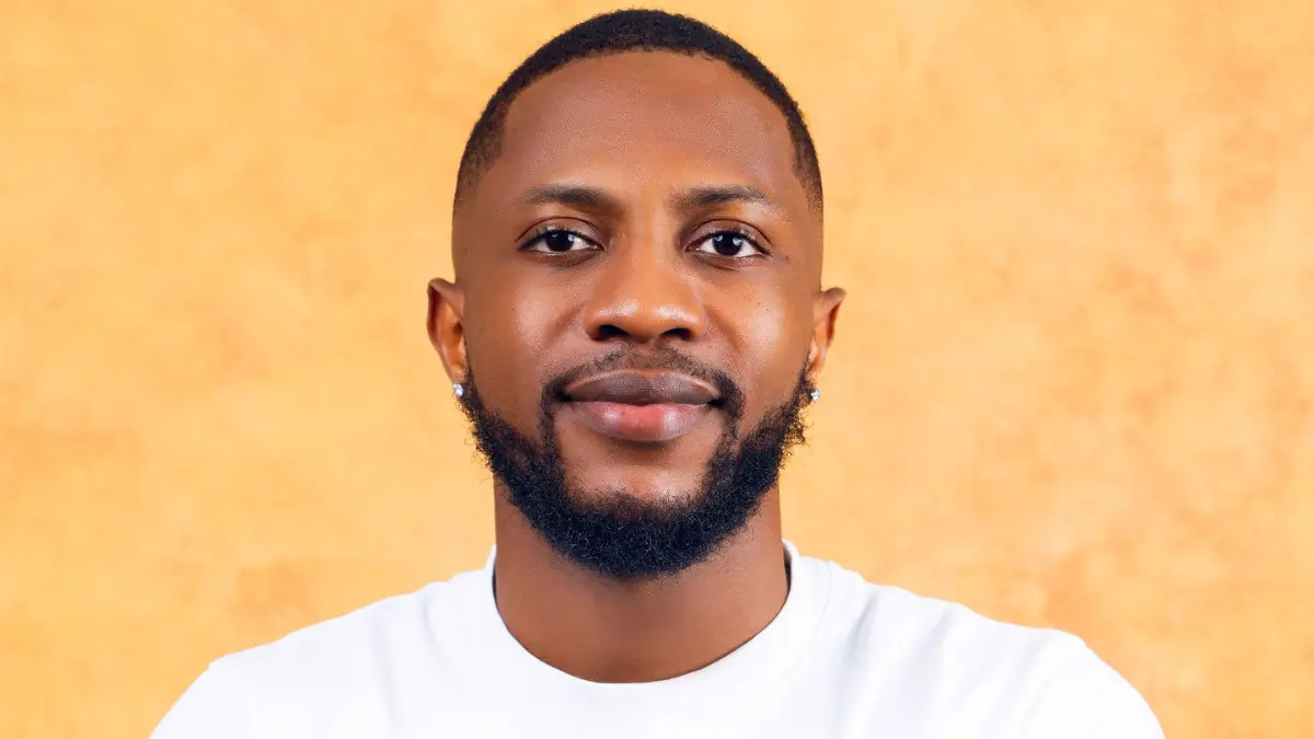 Kayvee - THEWILL NEWS MEDIA Close-up portrait of a man with a trimmed beard and earrings, wearing a white shirt, against an orange background.