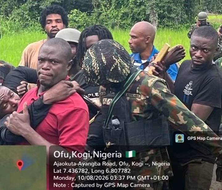 Kogi Kidnap Victims 1 - THEWILL NEWS MEDIA Group of men surrounding a restrained man in a red shirt in a grassy field; camouflage-wearing figures and bystanders visible; GPS map overlay reads Ofu, Kogi, Nigeria.