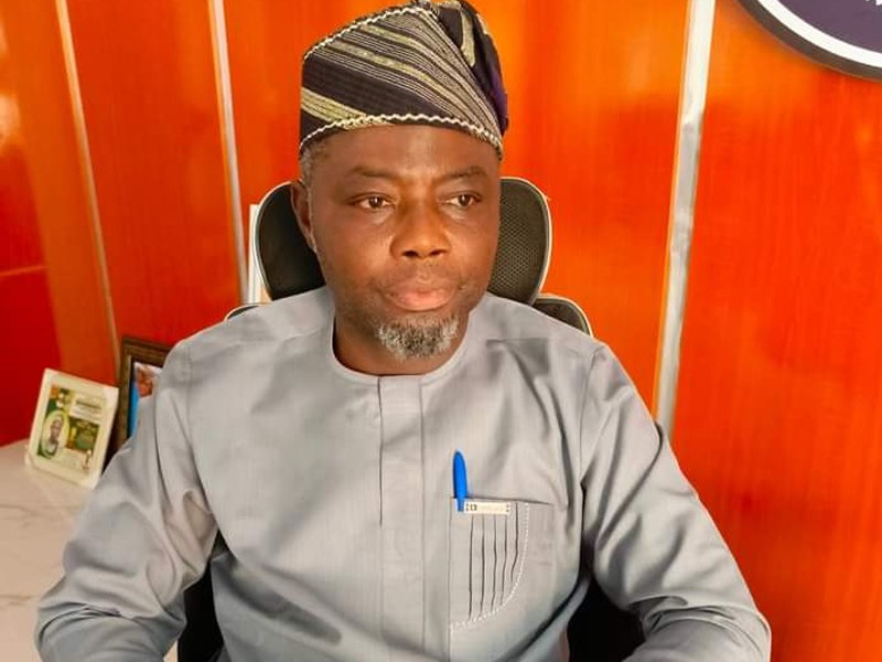 Kolapo Alimi - THEWILL NEWS MEDIA Professional man in a gray traditional outfit with a patterned cap sits at a desk in an orange-toned office; a blue pen is in his pocket.