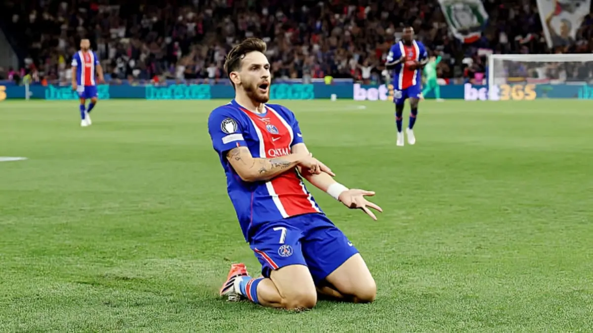 Kvaratskhelia - THEWILL NEWS MEDIA Soccer player in PSG blue-and-red kit kneels on the grass celebrating with outstretched arms as fans cheer in the background