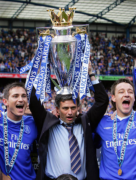 Lampard Terry and Mourinho - THEWILL NEWS MEDIA Three football players in blue kits celebrate on the pitch as the manager lifts a large gold trophy overhead, with blue ribbons streaming from it.