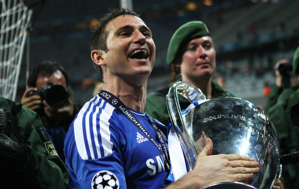 Lampard - THEWILL NEWS MEDIA Soccer player in a blue jersey lifts a large trophy in celebration, with photographers filming nearby in a stadium.