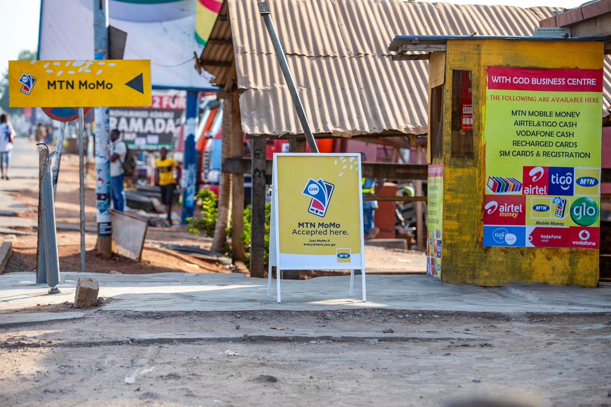 MTN MoMo Agent Infrastructure - THEWILL NEWS MEDIA Street market scene with bright MTN MoMo signs; yellow ad boards advertising mobile money services near a corrugated metal roof.