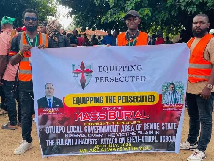 Members of a relief and advocacy group - THEWILL NEWS MEDIA Group of people in orange vests holding a large banner that reads 'Equipping the Persecuted' at a mass burial event in Nigeria, outdoors under trees.