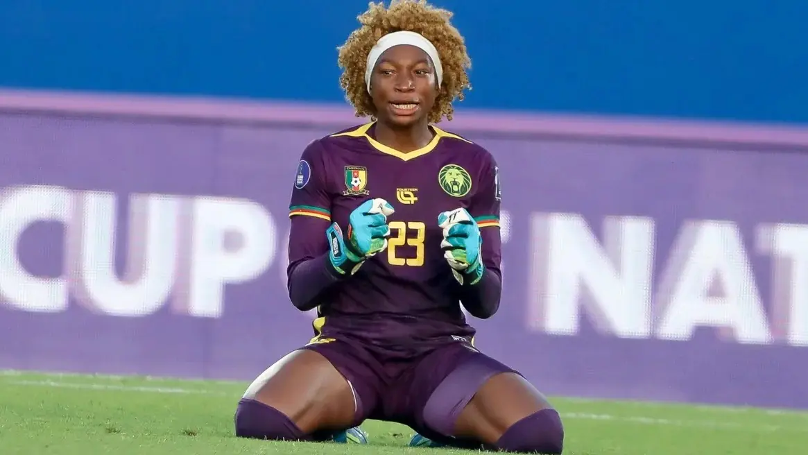 Michaely Bihina - THEWILL NEWS MEDIA Female soccer goalkeeper kneeling on the field, wearing a purple kit with number 23 and teal gloves, ready to defend the goal.