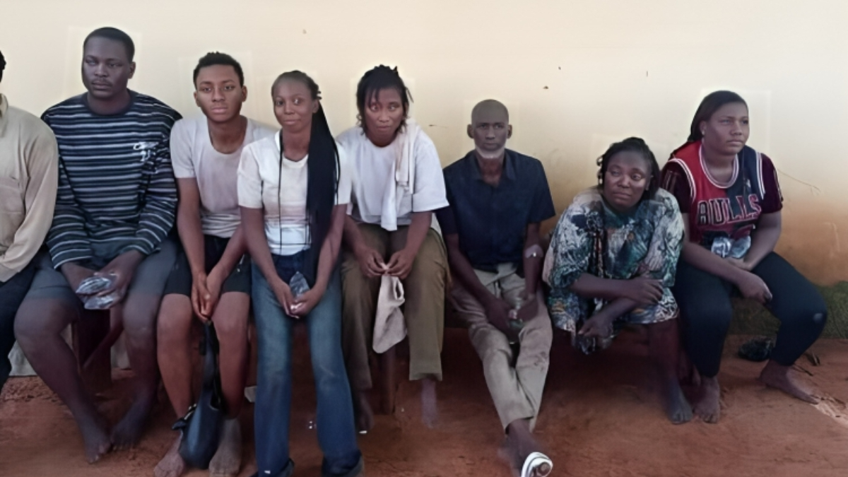 Military Rescues 15 NYSC Members - THEWILL NEWS MEDIA Group of eight people sitting on a bench against a pale wall, some holding bags, dressed in casual everyday clothes.