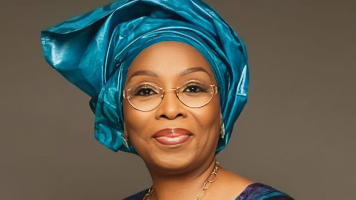 Modele Sarafa-Yusuf - THEWILL NEWS MEDIA Portrait of a smiling woman wearing a turquoise gele headwrap, round glasses, and gold jewelry against a neutral backdrop.