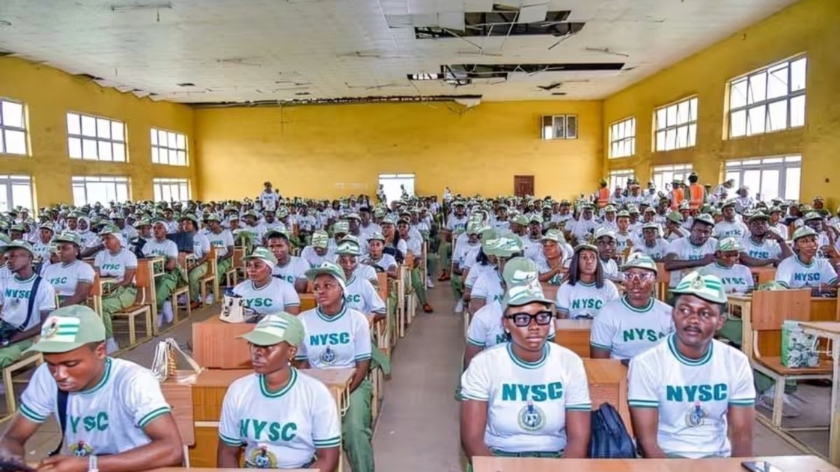 NYSC - THEWILL NEWS MEDIA Large hall filled with uniformed NYSC corps members seated at desks for a briefing or assembly in a yellow-walled room.