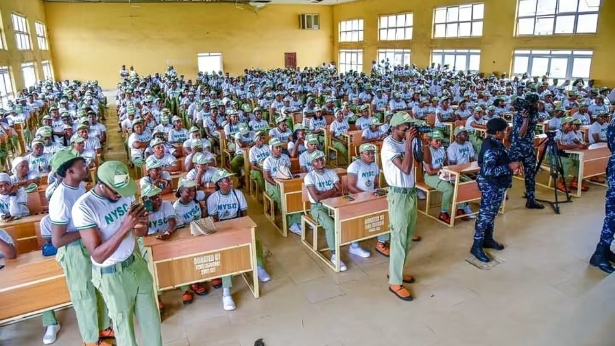 OSUN ELECTION USING NYSC - THEWILL NEWS MEDIA Large hall filled with hundreds of NYSC corps members in uniform white shirts and green pants seated at desks. A few standing and recording with cameras during the event.