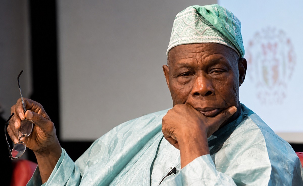 Olusegun Obasanjo - THEWILL NEWS MEDIA Older man in turquoise traditional clothing and a patterned cap rests his chin on his hand while holding glasses, deep in thought.