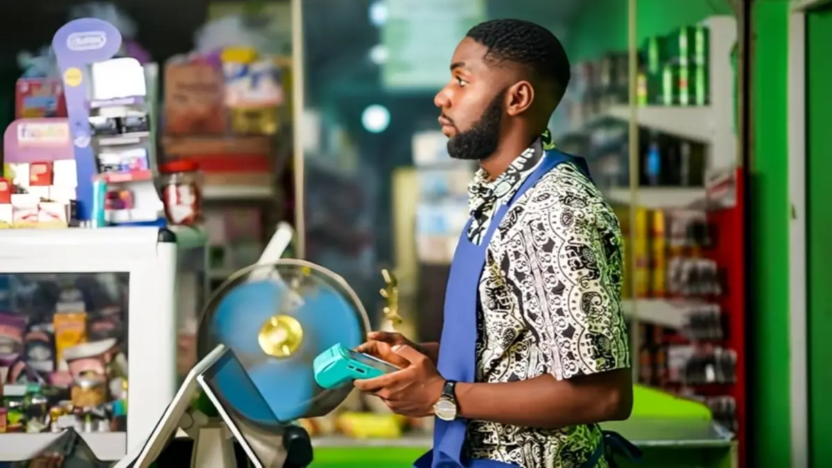Opay Merchant with pos - THEWILL NEWS MEDIA Shop attendant in a patterned shirt and blue apron handling a handheld card reader at a store counter.