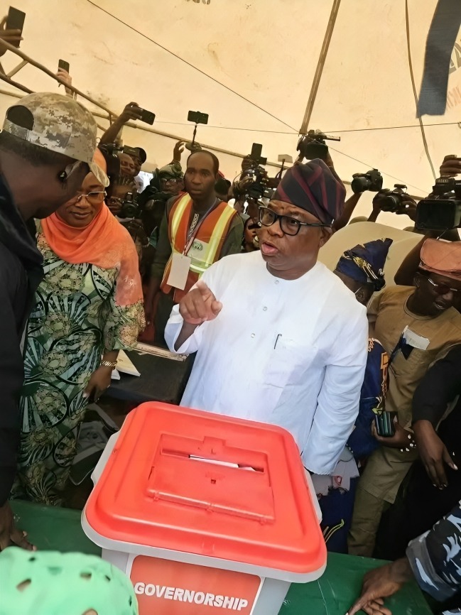 Osun APC governorship candidate Bola Oyebamiji -AMBO - THEWILL NEWS MEDIA Man in white traditional attire speaks near a red-lidded ballot box labeled'GOVERNORSHIP' as reporters film the voting event.