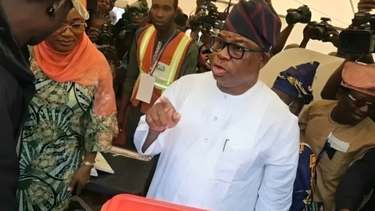 Osun APC governorship candidate Bola Oyebamiji AMBO - THEWILL NEWS MEDIA Man wearing a white traditional outfit and glasses gestures while surrounded by photographers and onlookers in a crowded scene.