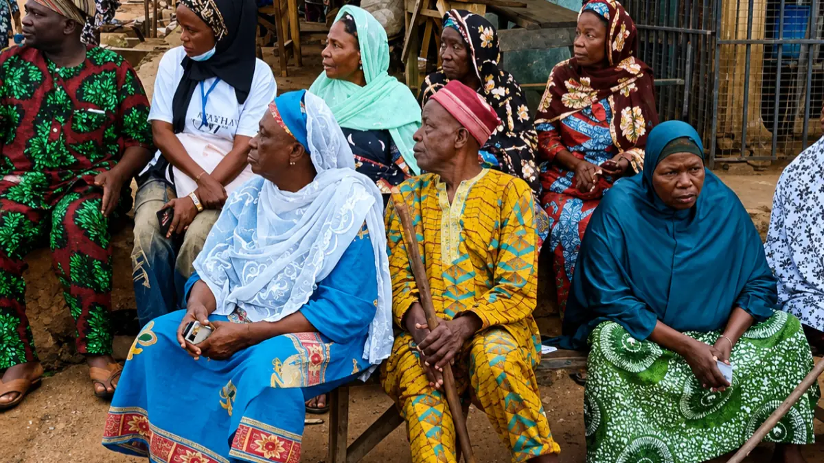 Osun voters - THEWILL NEWS MEDIA Group of West African adults sitting outdoors, wearing vibrant traditional clothing, watching something off-camera, suggesting a community gathering.