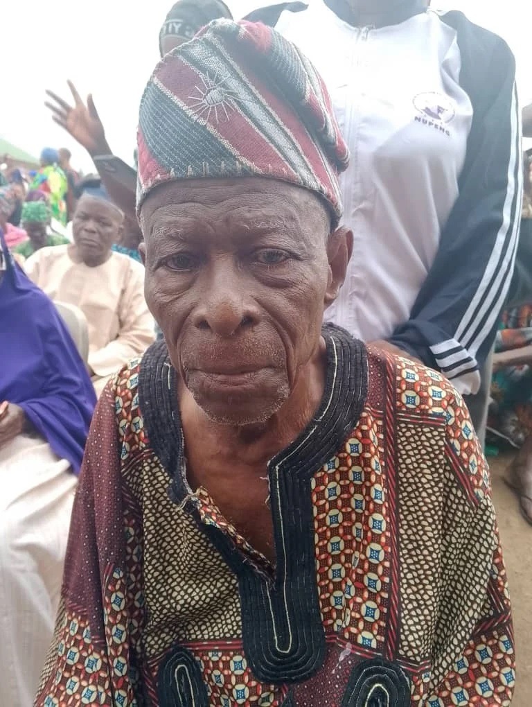 Pa Odejide Mosunmoye - THEWILL NEWS MEDIA Older man in a colorful traditional patterned robe and a patterned cap sits at an outdoor event, with people in the background.